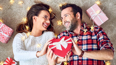 Glückliches Pärchen hält unterm Weihnachtsbaum ein Herzgeschenk in der Hand